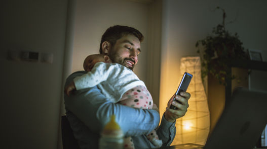 En man står upp med en liten bebis vilande på axeln. I sin lediga hand håller mannen en mobiltelefon.