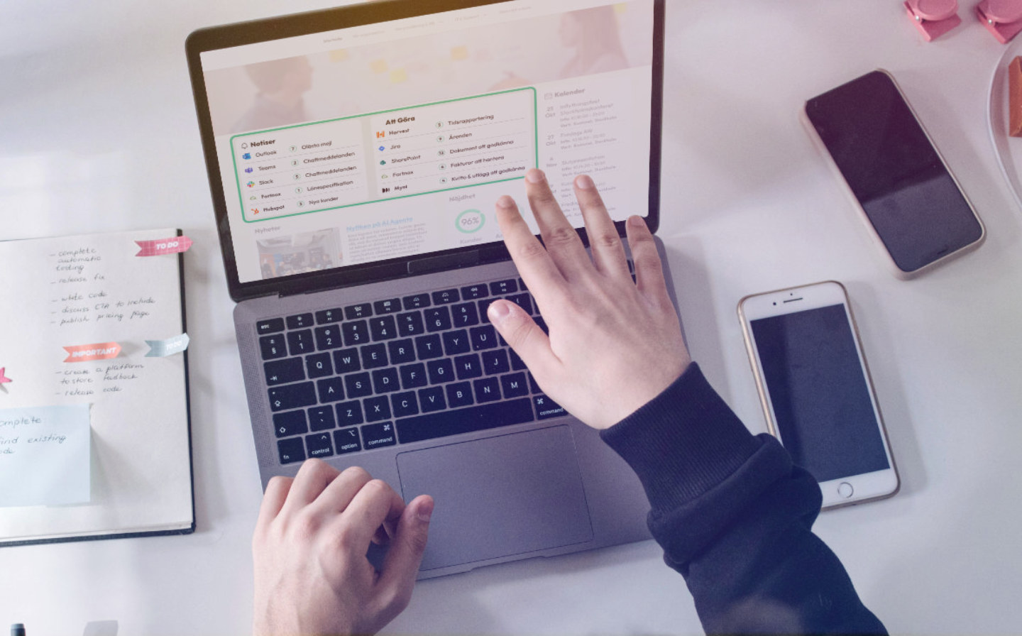 En person arbetar vid en b&auml;rbar dator p&aring; ett skrivbord. P&aring; sk&auml;rmen visas ett digitalt kontrollpanel- eller intran&auml;tsgr&auml;nssnitt med en att-g&ouml;ra-lista och olika systemikoner. P&aring; skrivbordet ligger &auml;ven en mobiltelefon och anteckningar.